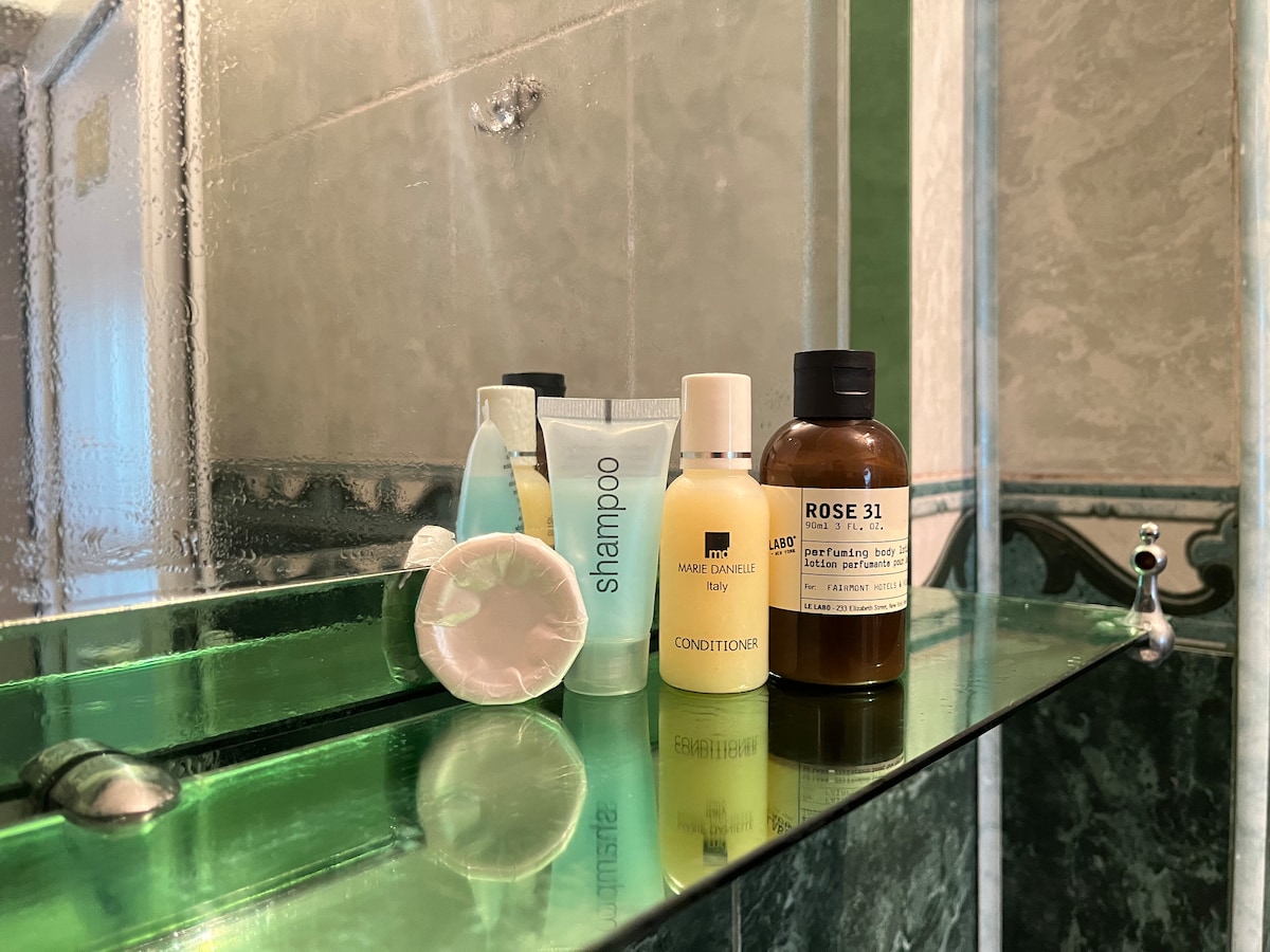 A selection of personal care items is displayed on a glass shelf. The arrangement includes shampoo, conditioner, and a small facial product, all resting against a backdrop of textured green tiles and a wide mirror reflecting the products.