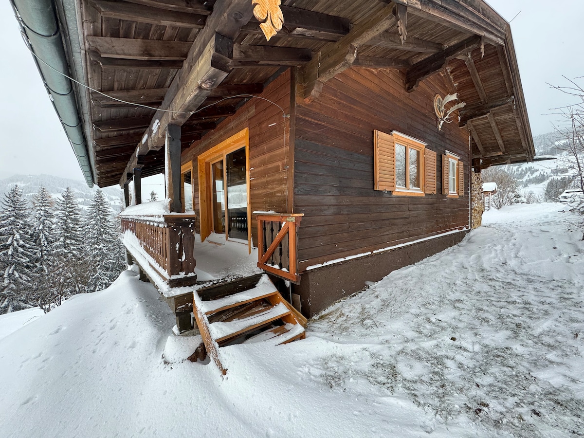 The exterior of the rustic wooden cabin is framed by a snowy landscape, featuring a welcoming entrance with a wooden railing and staircase. Large windows provide views of the surrounding winter scenery, while snow-covered trees can be seen in the distance.