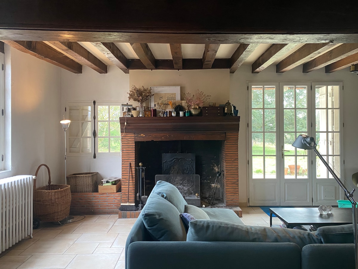 A living room features exposed wooden beams and a large fireplace, enhancing the rustic charm. Natural light streams through multiple glass doors, offering views of the greenery outside. A comfortable sofa and minimalist decor contribute to a calming atmosphere.