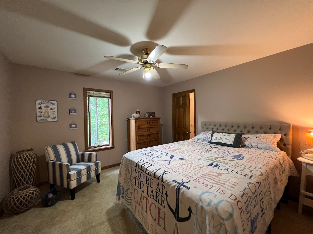 A nautical-themed bedroom features a spacious bed adorned with themed linens. A window provides a view of the outdoors, while a cozy armchair is positioned nearby. A dresser is located against the wall, and soft lighting creates a comfortable environment.