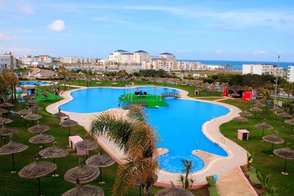 The image captures a wide view of a large pool surrounded by meticulously maintained green lawns and palm trees. Numerous thatched umbrellas provide shaded areas. In the distance, buildings and the coastline are visible under a clear blue sky, creating a serene outdoor space.