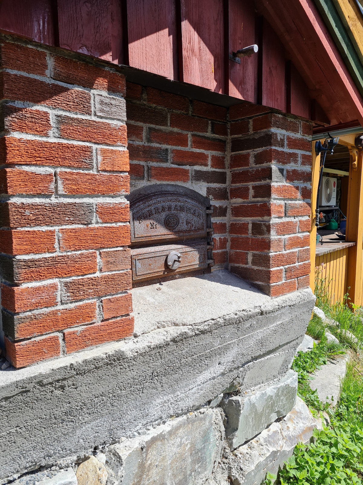 An antique mailbox made of cast iron is mounted on a stone and brick base. Red bricks create a textured backdrop, while nearby greenery adds a natural touch to the scene. The mailbox's decorative details are visible, enhancing its rustic charm.