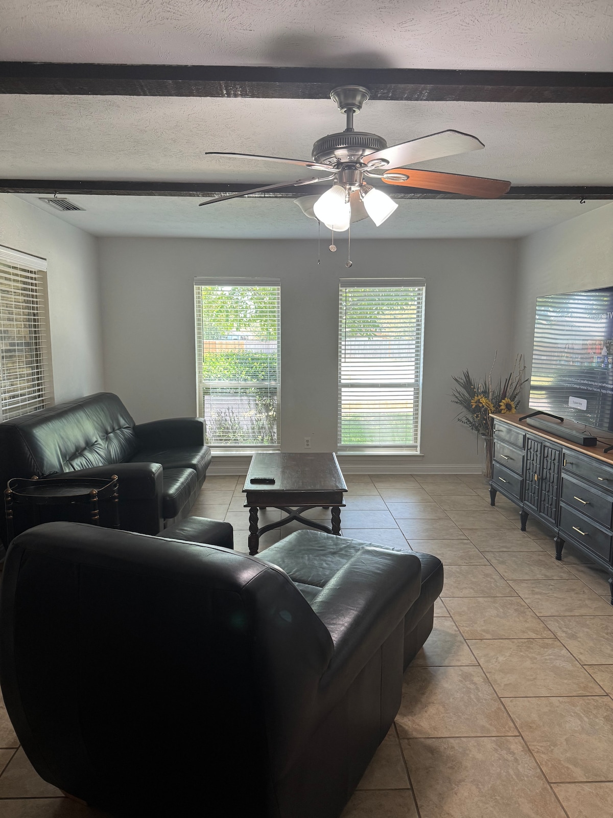 A living area is highlighted by two black leather sofas arranged around a wooden coffee table. Natural light filters through multiple windows adorned with horizontal blinds, creating a bright atmosphere. A decorative console stands against the wall, complemented by a subtle floral arrangement.