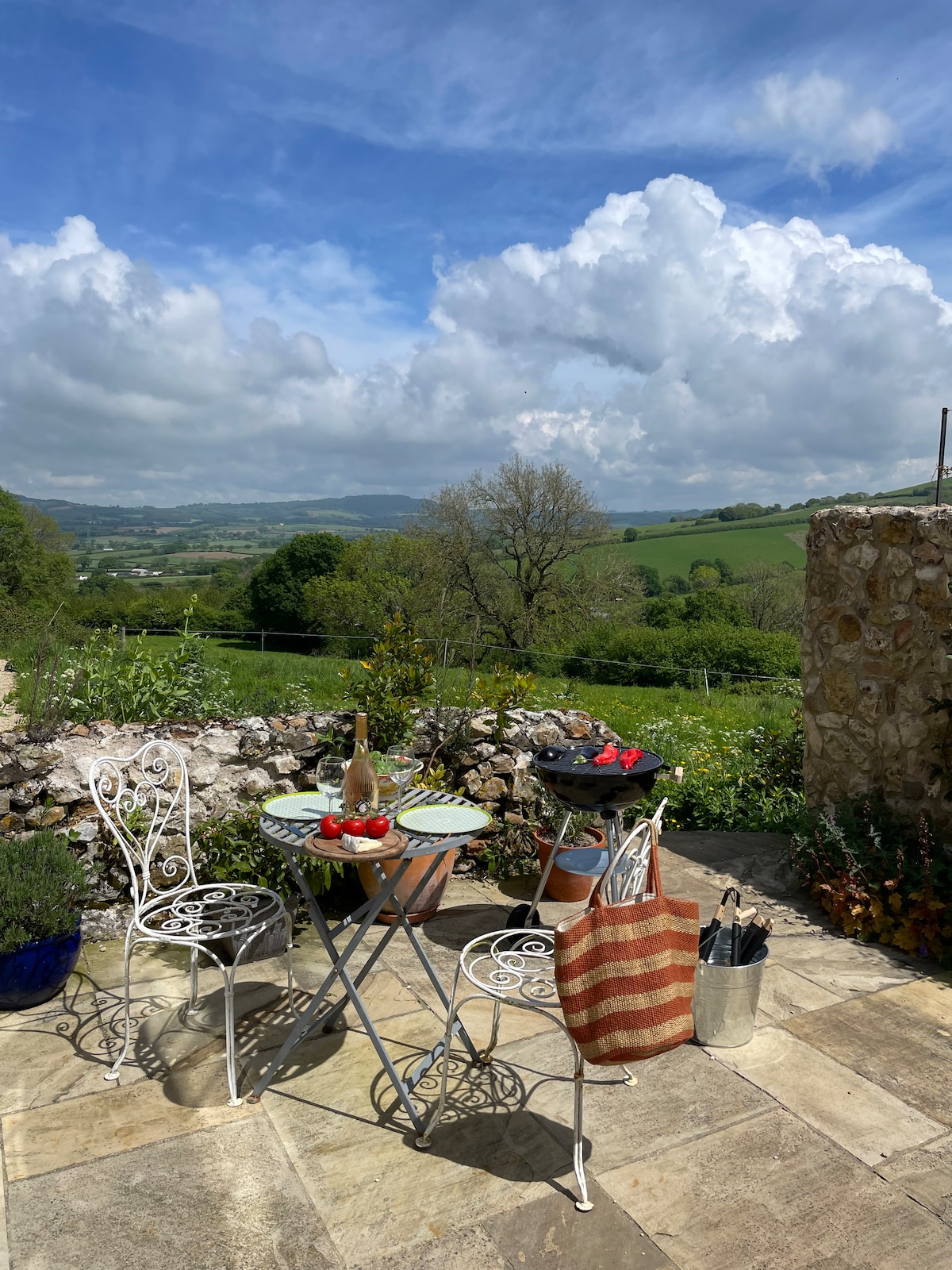 A quaint private terrace is shown, featuring a round table with two chairs, set with fresh produce and a bottle. A BBQ stands nearby, surrounded by lush greenery and a natural stone wall. The expansive view of rolling hills and bright blue sky creates a serene backdrop.