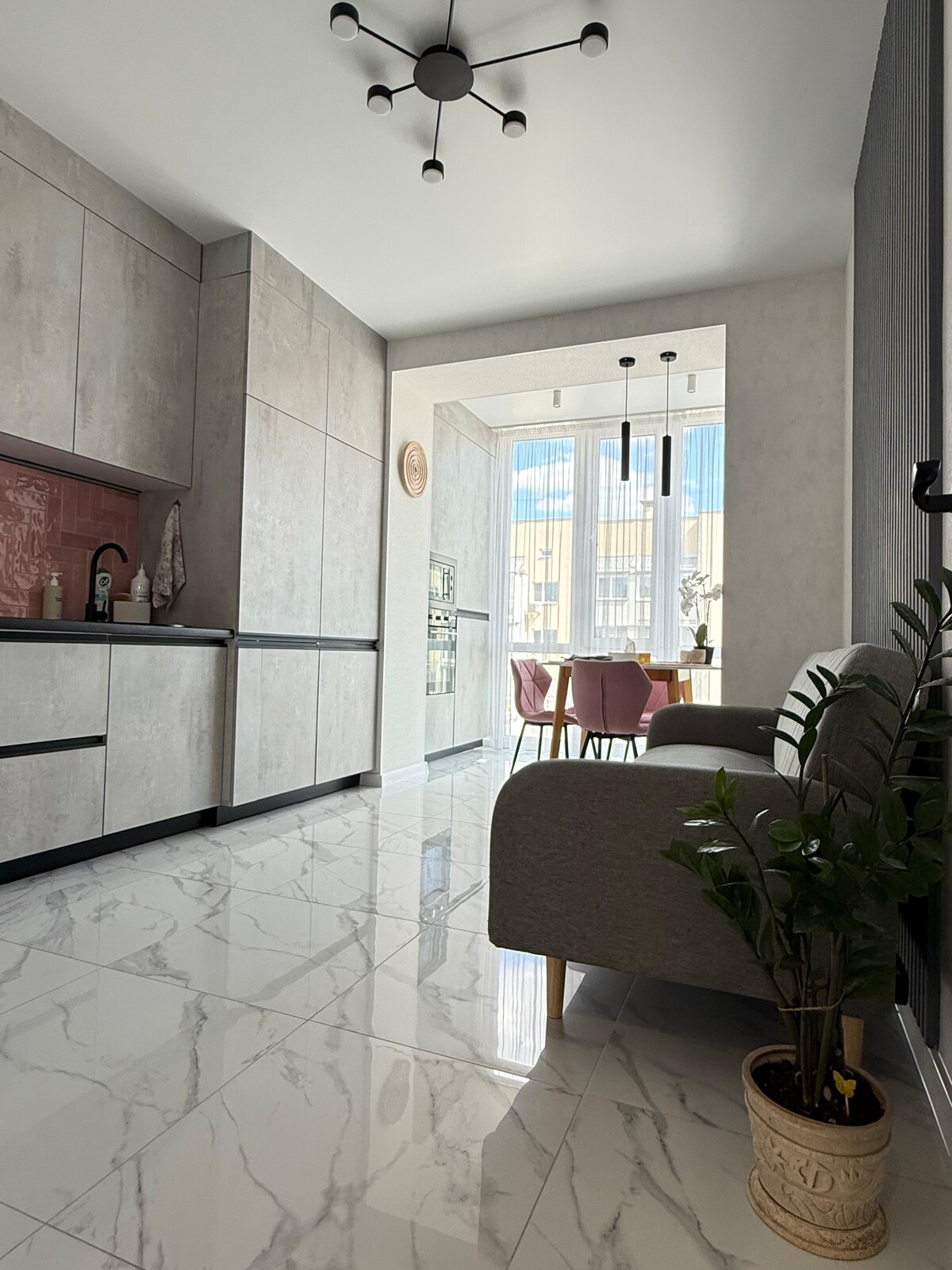 A modern kitchen features sleek gray cabinetry and a minimalist design. Natural light fills the dining area, where a table accompanied by pink chairs is visible. A cozy seating area is positioned with a small plant on the floor, enhancing the functional layout.