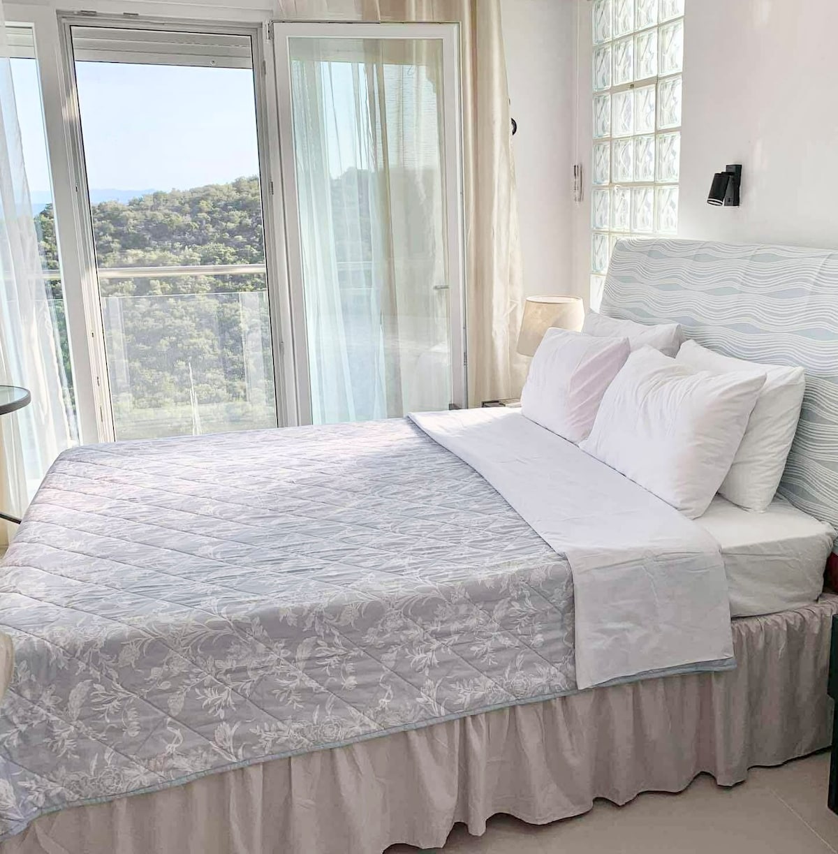 A spacious bed is dressed in light linens, accompanied by multiple pillows. Natural light is filtered through sheer curtains, enhancing the serene environment. A glass door leads to a balcony with views of the surrounding landscape, creating a connection to the outdoors.