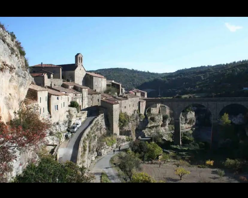A serene village is nestled among rolling hills, characterized by stone buildings and a prominent church tower. An archway bridge spans a valley, connecting pathways lined with trees and open spaces, enhancing the tranquil ambiance of the landscape.