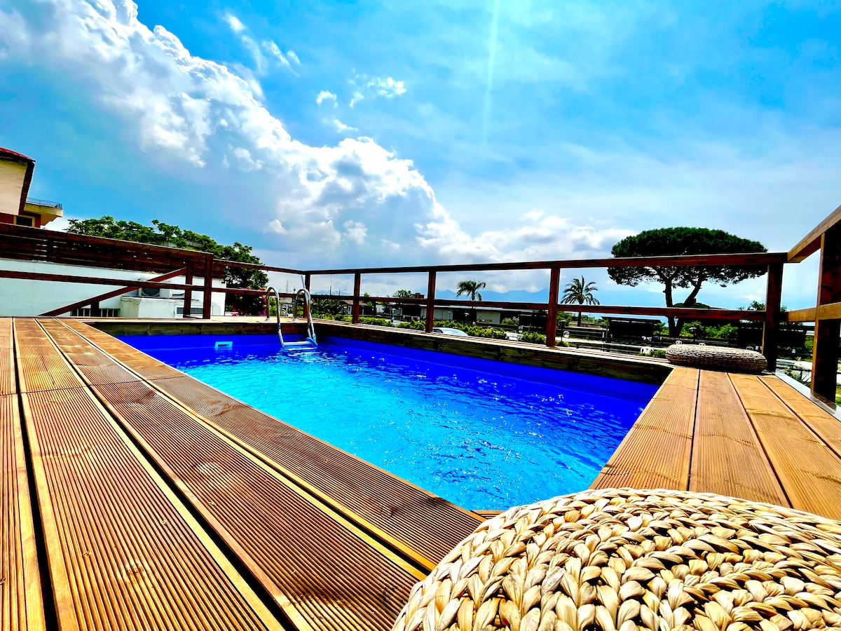 A luxurious wooden swimming pool is framed by a deck, with inviting blue water reflecting the sky. Palm trees are visible in the background, set against a backdrop of fluffy clouds, contributing to a serene outdoor environment.