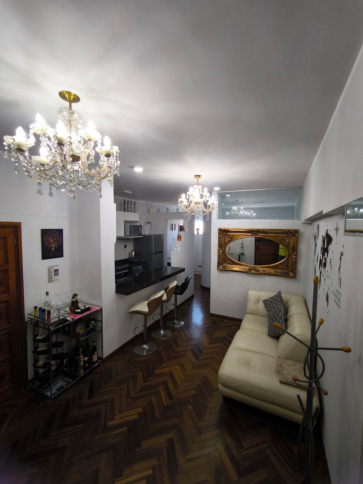 A stylish living area showcases modern bar stools next to an open kitchen. Elegant chandeliers provide soft lighting, while a decorative mirror enhances the spacious feel. A light-colored couch invites relaxation, and polished wooden flooring adds warmth to the ambiance.