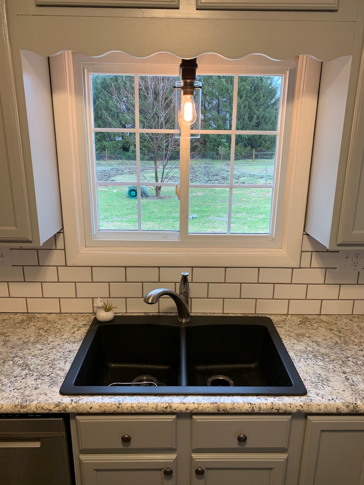 A modern kitchen features a double-basin black sink underneath a grid-patterned window. Natural light illuminates the space, highlighting the light gray cabinetry and white subway tile backsplash. A single pendant light hangs above the sink, enhancing the practical workspace.