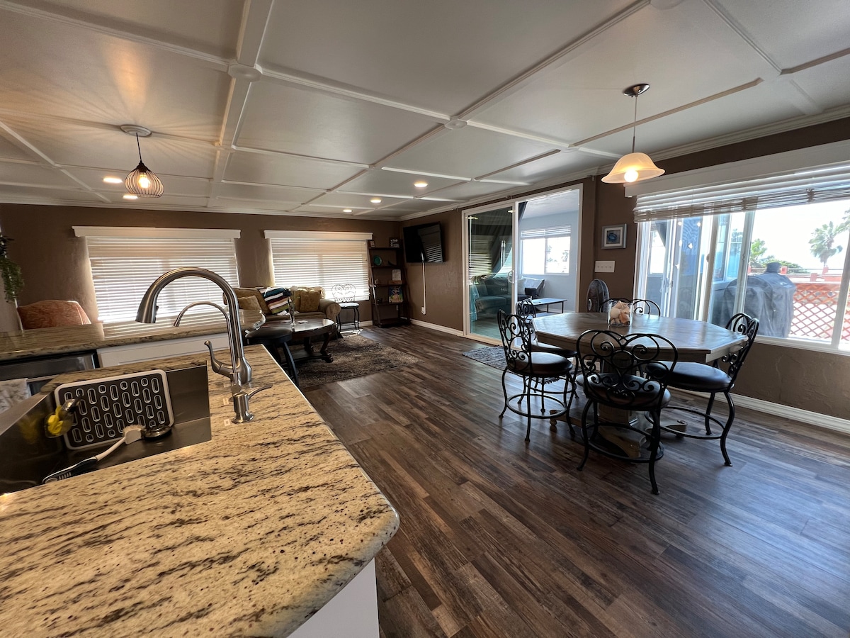 An open-concept living space is displayed, featuring a kitchen with a granite countertop and a dining area with a round table surrounded by black metal chairs. Large windows provide natural light, revealing a comfortable sofa and decor in the background.