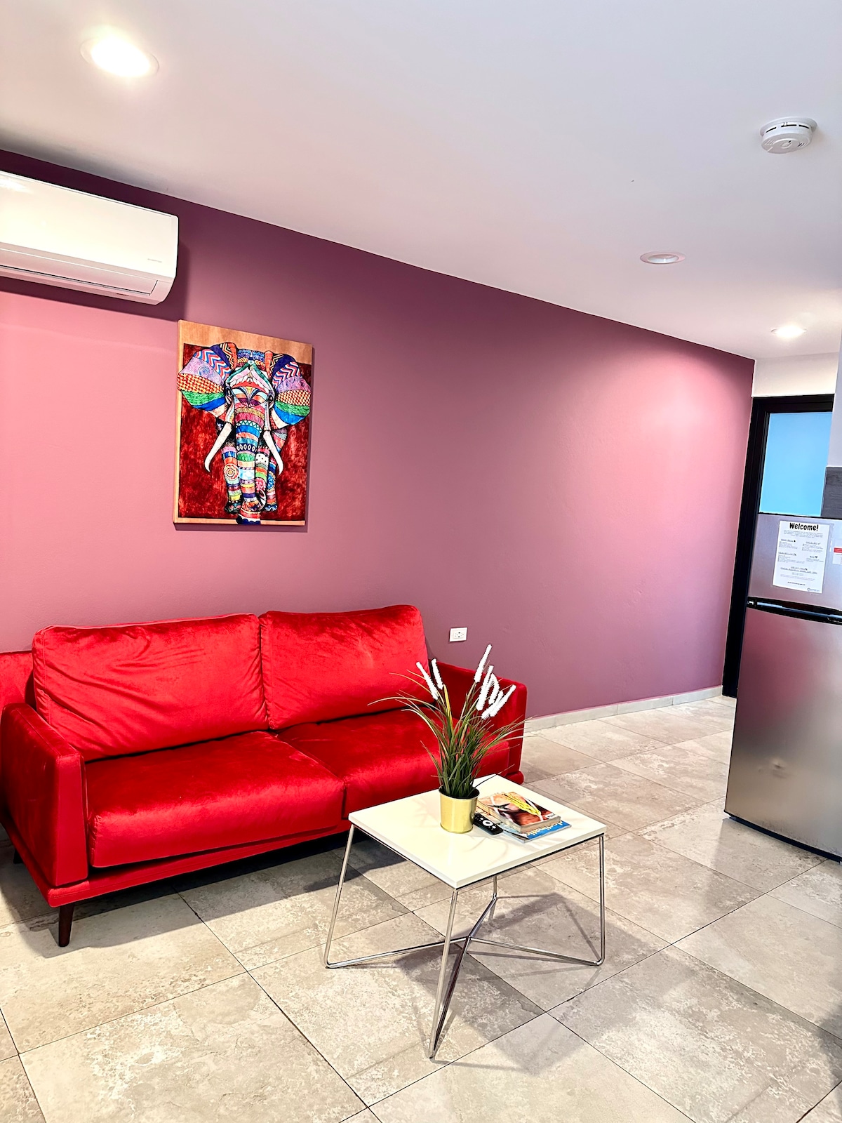 A vibrant red couch is positioned against a purple wall, complemented by a colorful wall art piece featuring an elephant. A small glass table holds a plant and magazines. The room has a modern, open layout, with light-colored tile flooring and a refrigerator visible in the background.