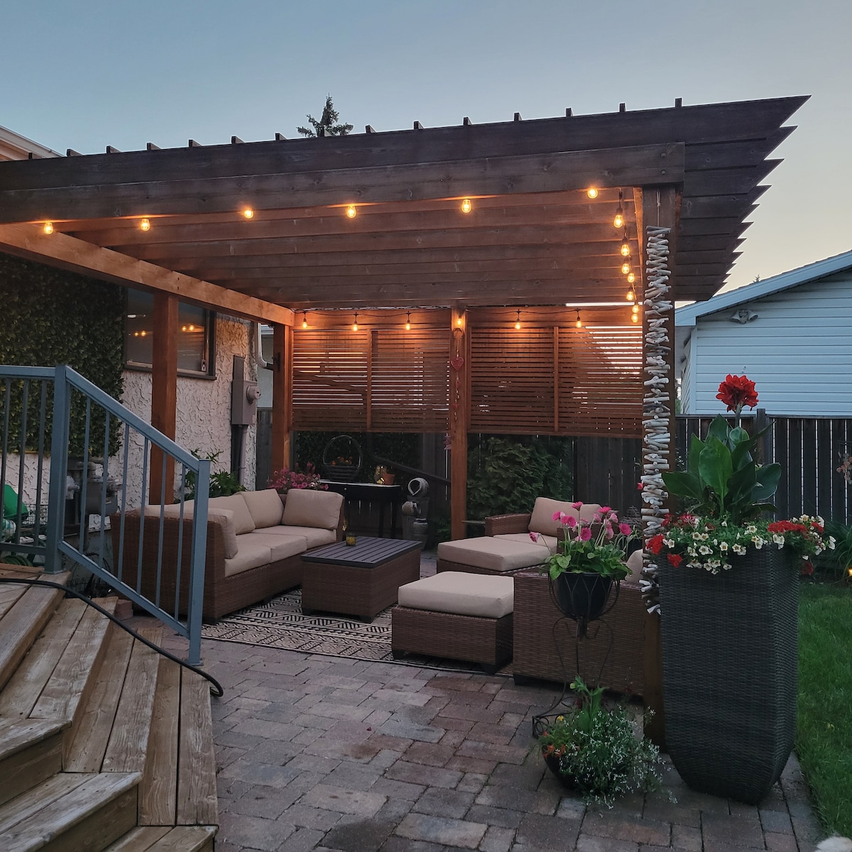 A covered outdoor patio area features comfortable seating arranged around a small table. Soft string lights illuminate the space, creating an inviting atmosphere. Potted flowers add pops of color, while the adjacent landscaped greenery enhances the outdoor experience.