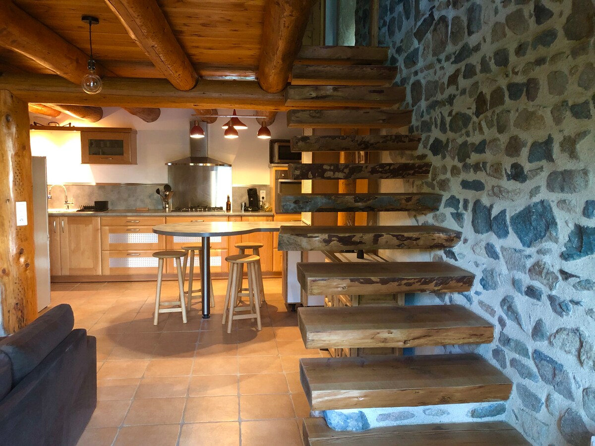 A rustic interior showcases wooden beams and stone walls. A staircase made of wooden steps leads to the upper level. A round table and two bar stools are positioned near a modern kitchen equipped with essential appliances. Warm natural light fills the space.