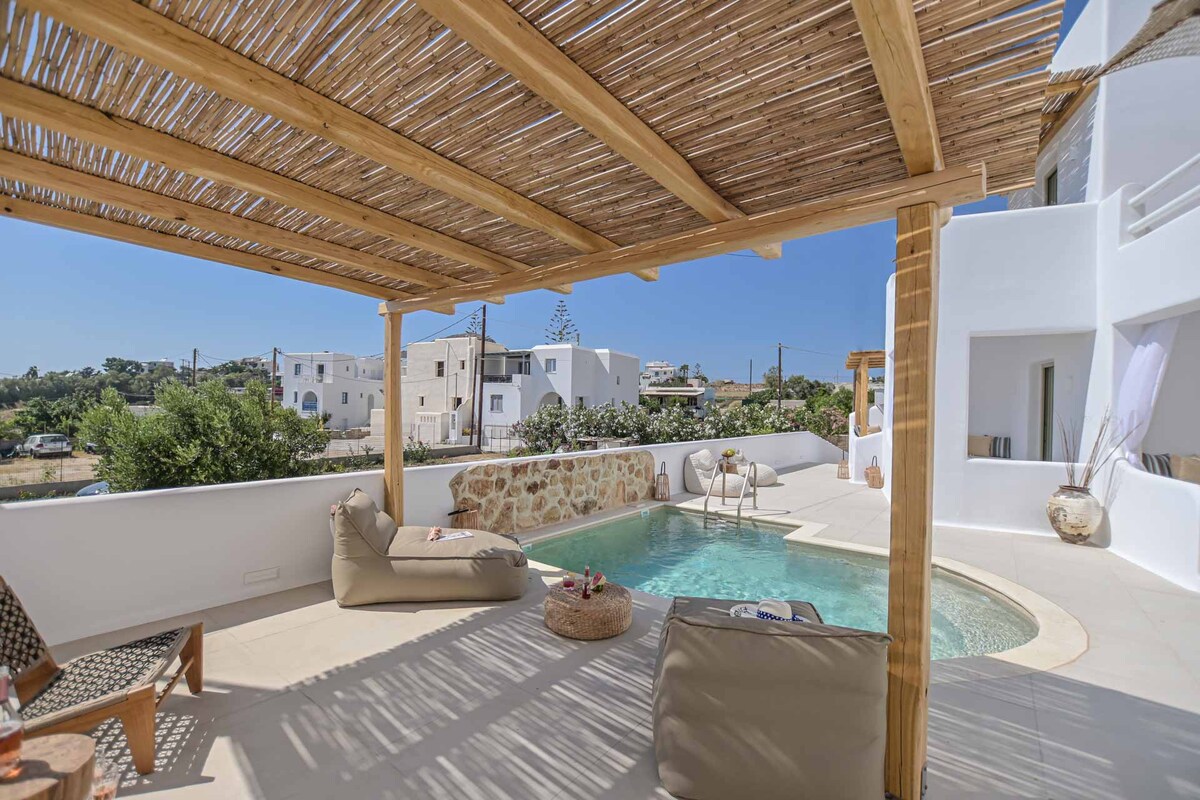 A shaded seating area is positioned next to a serene pool, surrounded by a mix of greenery and architecture. Natural elements like wood and stone are present, contributing to the outdoor lounge atmosphere, while clear skies can be seen in the background.