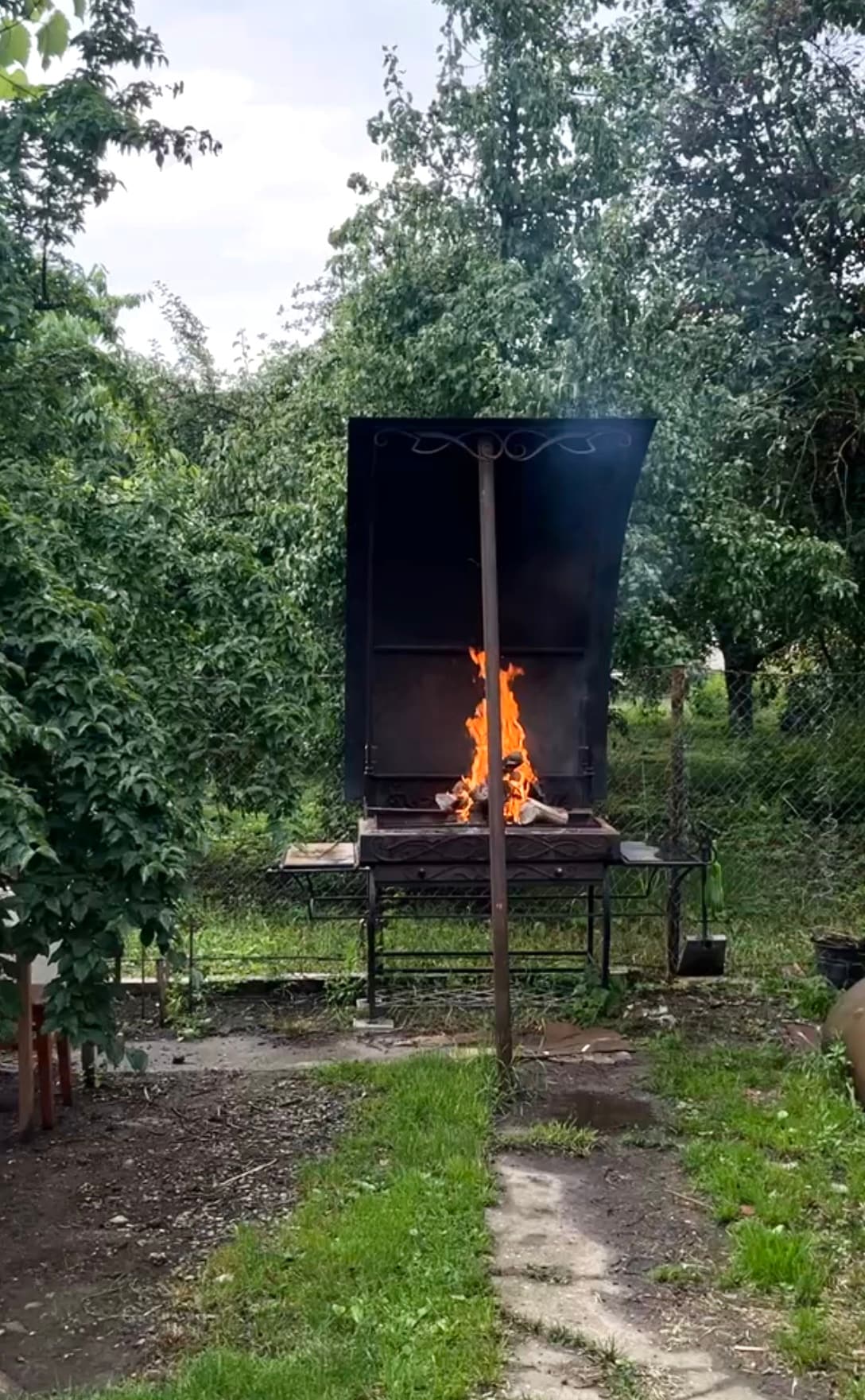An outdoor cooking area is visible, featuring a barbecue grill with flames rising from it. Surrounding greenery creates a natural backdrop, enhancing the calm setting. The ground is partly covered with grass, and a rustic structure supports the grill.