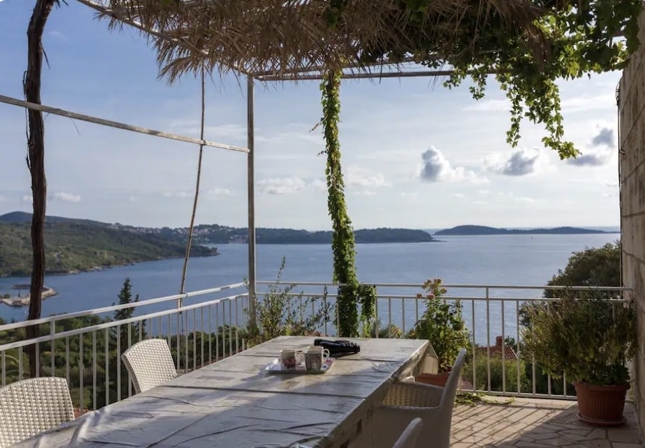 A balcony area is presented with a large table surrounded by several light-colored chairs. Lush greenery and a straw canopy provide partial shade. The view features calm waters and distant hills, contributing to a serene outdoor setting.
