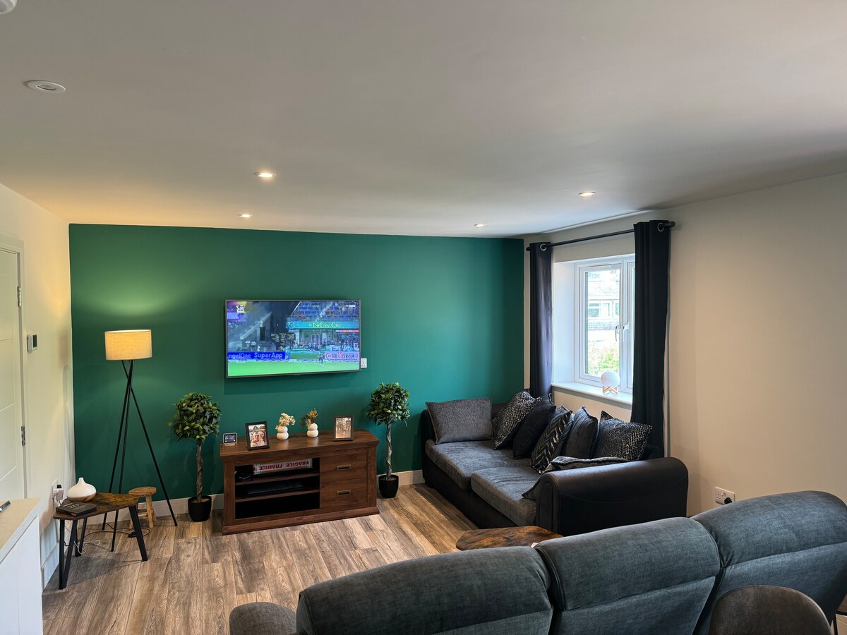 A modern living area is highlighted by a large television mounted on the wall above a wooden media console. Soft seating is provided by a dark grey sectional sofa, complemented by decorative plants and a standing lamp. Natural light enters through a nearby window adorned with dark curtains.