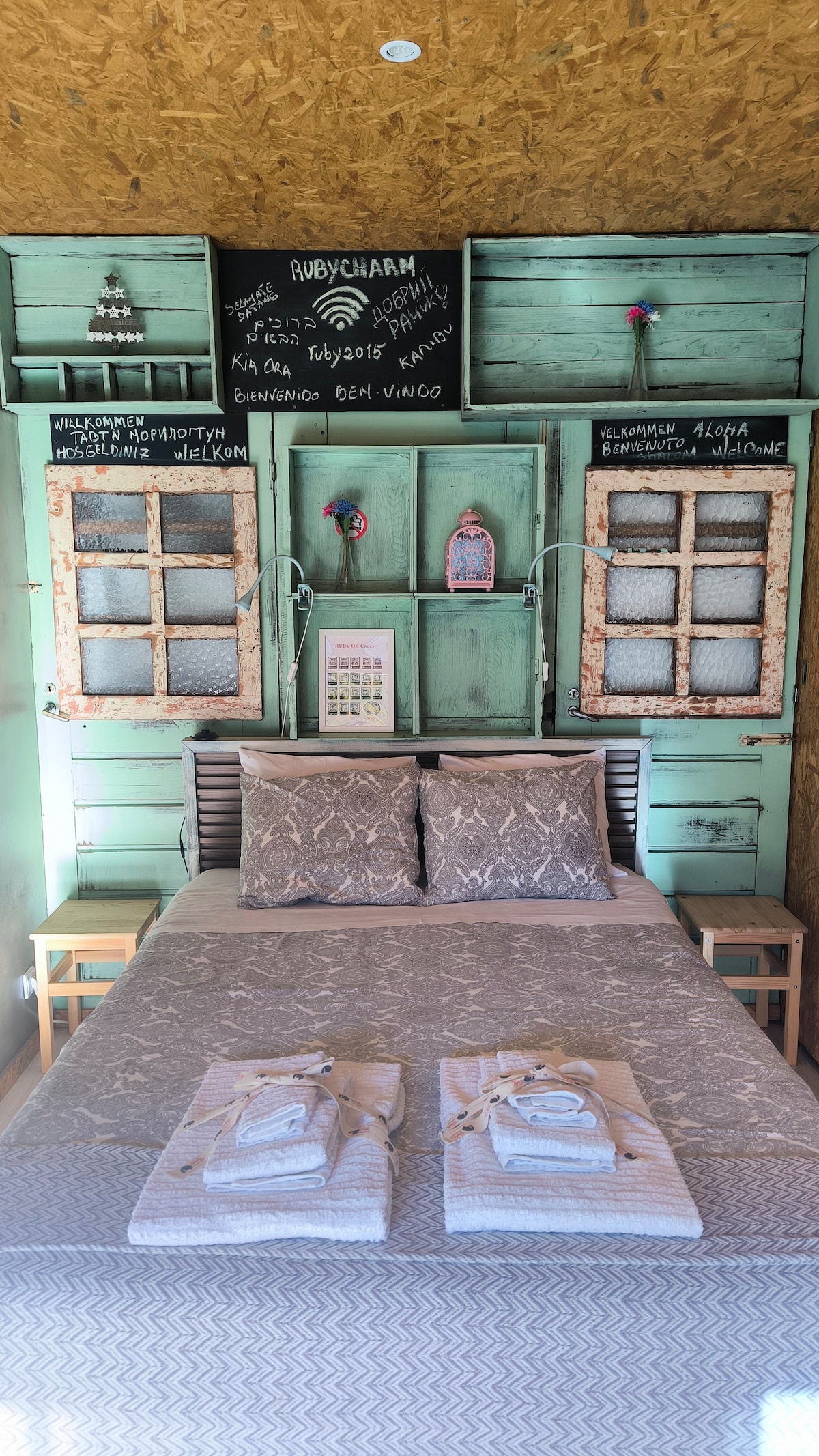 The sleeping area features a spacious bed with light-colored linens and decorative pillows. Behind the bed, rustic wooden panels in a soft green color frame several small windows, creating a charming backdrop. Two small nightstands accompany the bed, adding functional elegance.