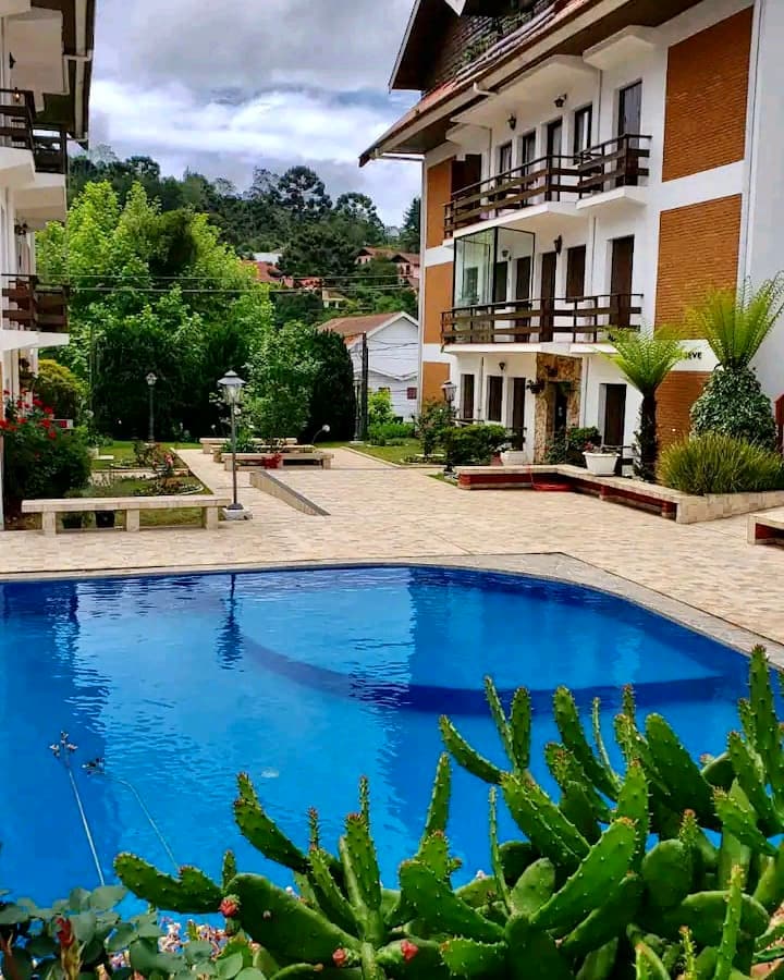 A serene outdoor area features a sparkling pool surrounded by well-maintained gardens and greenery. The multi-story building showcases balconies with wooden railings, offering a welcoming atmosphere. Pathways and landscaping enhance the overall ambiance, ensuring a relaxing environment for guests.
