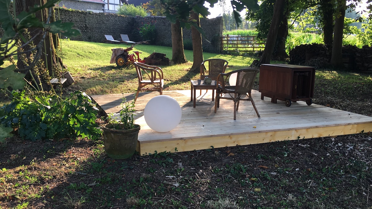 A wooden deck is situated in a grassy area surrounded by trees. A small round light is placed on the deck, accompanied by a few woven chairs and a wooden table. In the background, sun loungers are partially visible, creating a serene outdoor space.