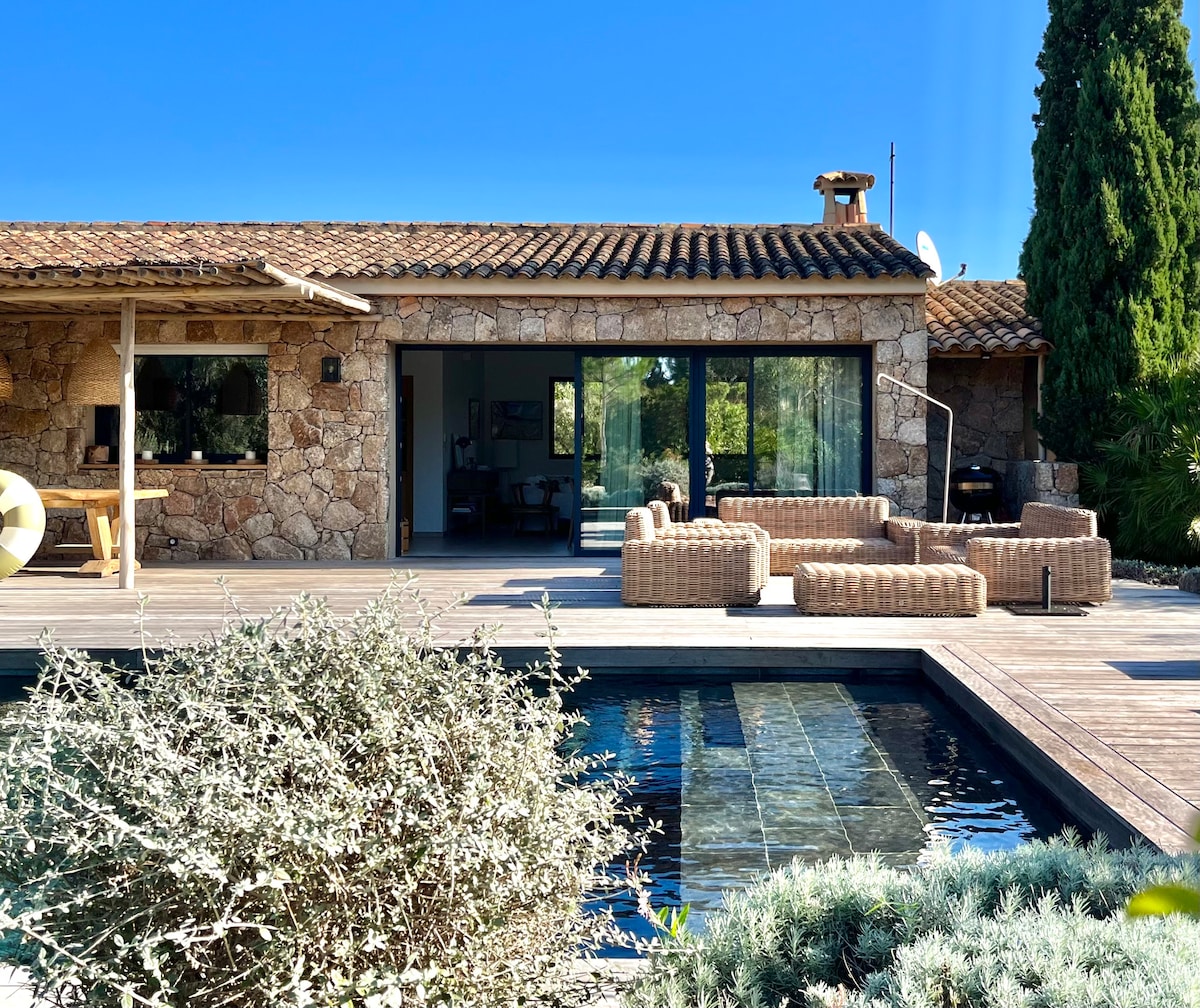 The exterior of a villa features natural stone walls and a terracotta tiled roof. A spacious terrace surrounds a clear swimming pool, while comfortable wicker seating is positioned nearby. Lush greenery complements the serene atmosphere, showcasing a blend of outdoor relaxation and elegant design.