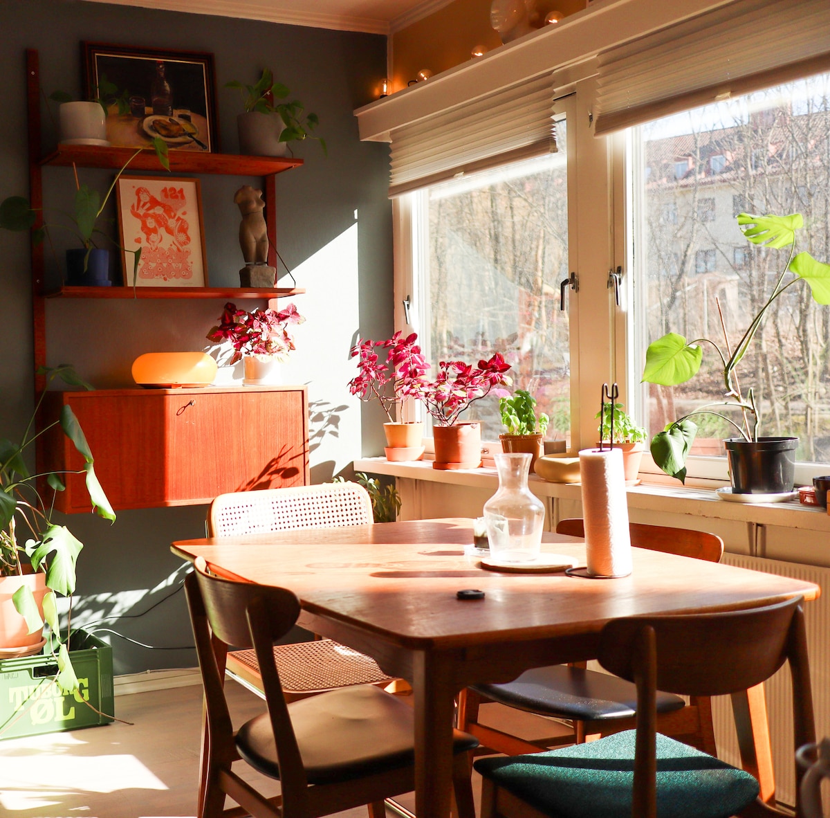 A sunlit dining area showcases a wooden table surrounded by four chairs. A collection of potted plants adorns the windowsill, adding natural greenery to the space. The wall features a shelf with art and decorative items, creating a cozy and inviting atmosphere.