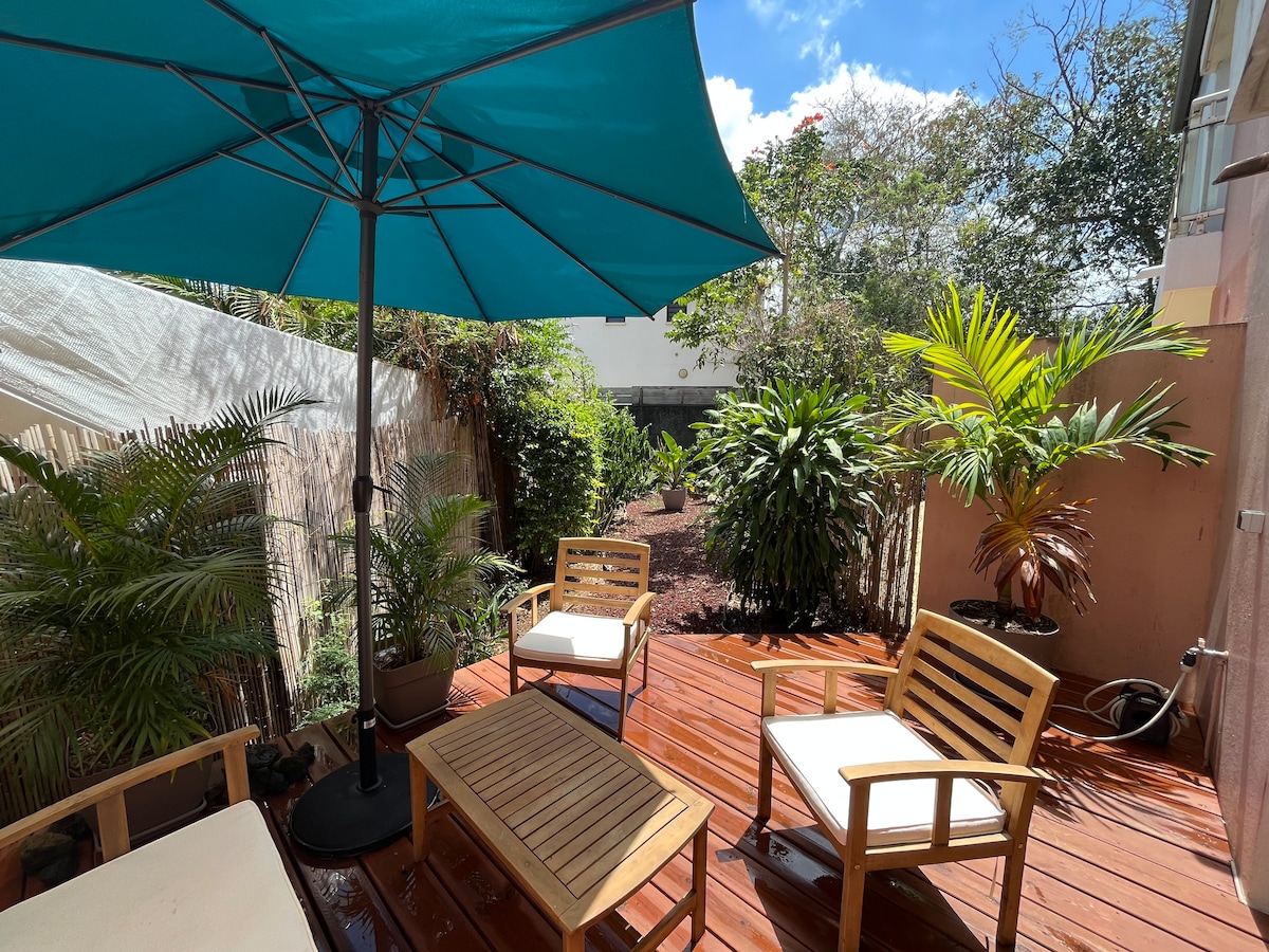 An outdoor terrace is showcased, featuring wooden decking surrounded by lush greenery. A large, teal parasol provides shade over two wooden chairs and a small table, creating a comfortable space to relax. Sunlight filters through the trees, illuminating the area.