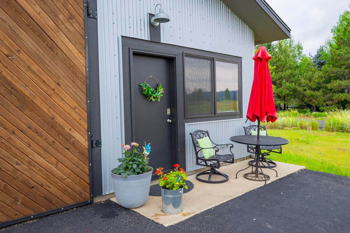 The entrance to the studio features a black door adorned with a decorative wreath. A small round table with two chairs is positioned beside it, covered by a red umbrella. Potted plants with colorful flowers add charm to the welcoming outdoor space.
