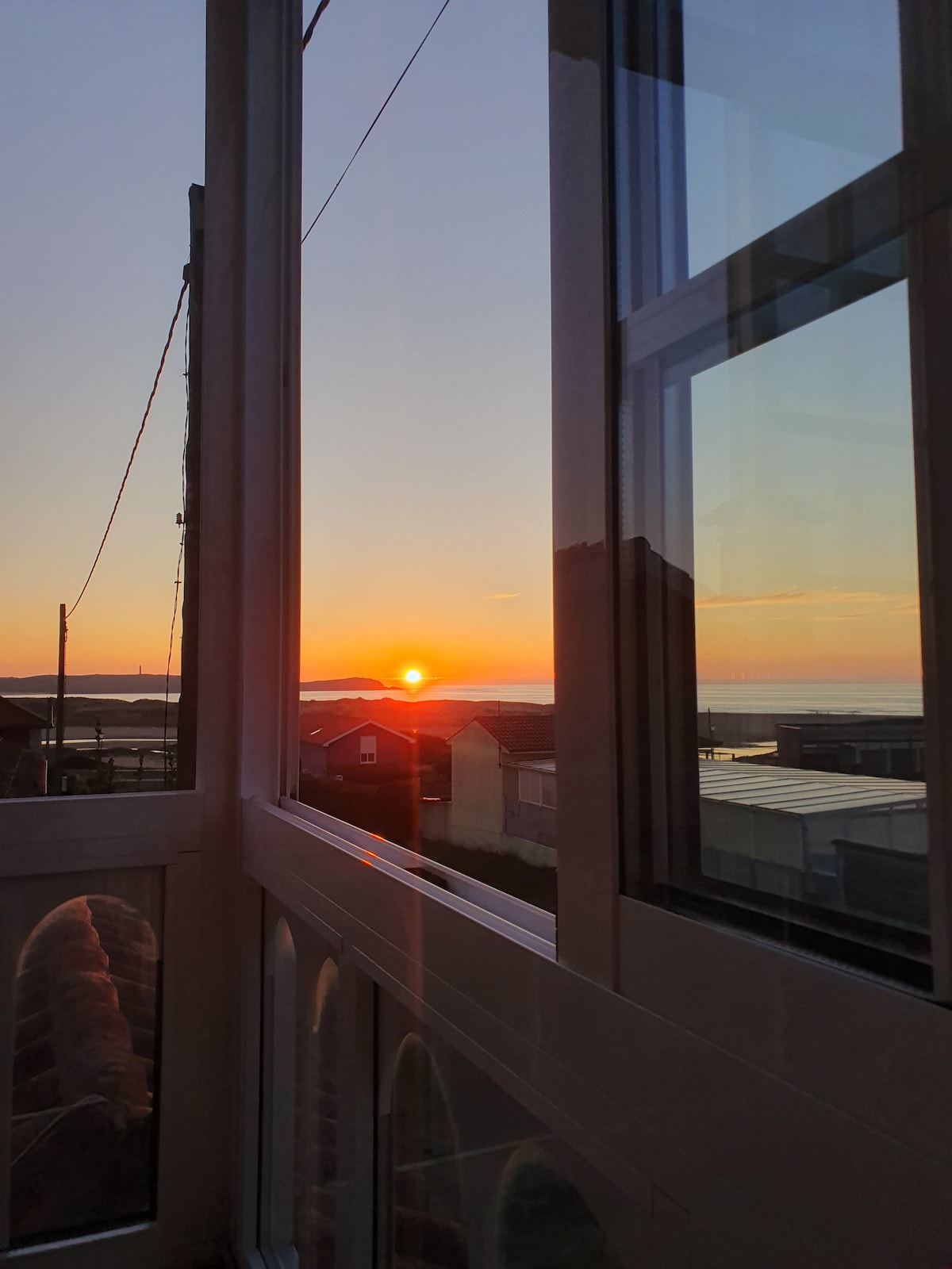 A stunning view is captured through large glass windows, showcasing a vibrant sunset over the ocean. The warm hues of orange and pink paint the sky as the sun dips below the horizon, while the rooftops of nearby houses are silhouetted against the backdrop.