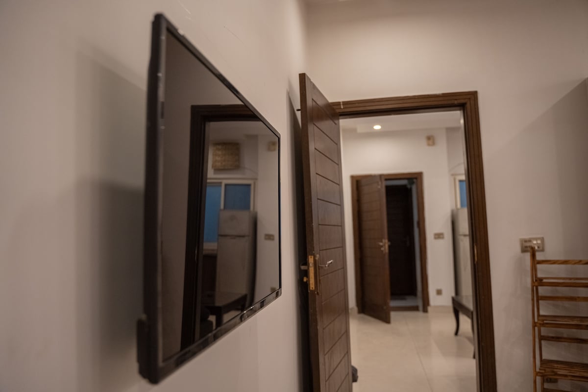 A flat-screen TV is mounted on the wall near a wooden door, leading to a softly lit hallway. The doorway opens to a glimpse of additional rooms, featuring light-colored walls and neutral-toned flooring. Natural light filters through several nearby windows.
