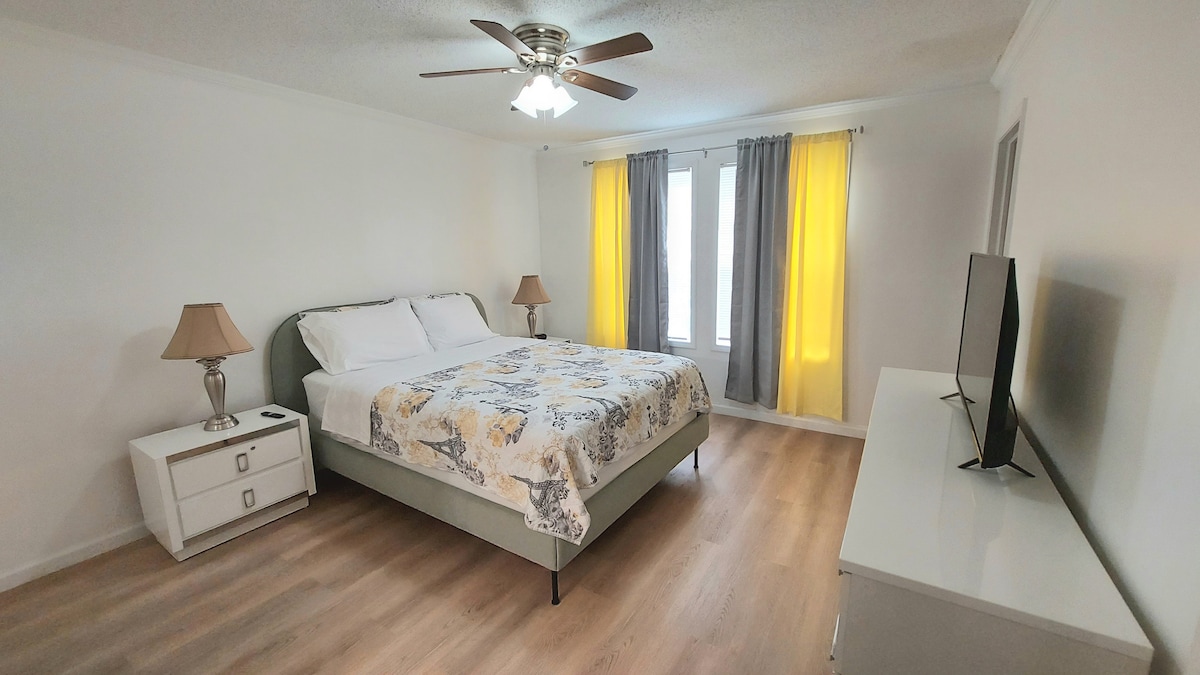 A spacious bedroom is presented with a large bed covered in a floral-patterned bedspread. Two nightstands with lamps flank the bed. Natural light filters through the sheer curtains, with one window dressed in yellow drapes. A television is positioned on a low white dresser.