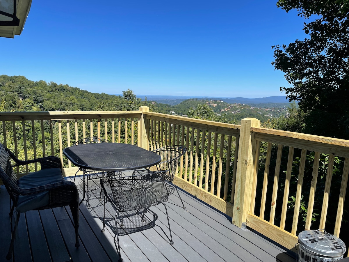 An outdoor deck is displayed with a round black table surrounded by wicker chairs. The railing offers an uninterrupted view of the surrounding mountains and blue sky, providing a serene backdrop for relaxation.