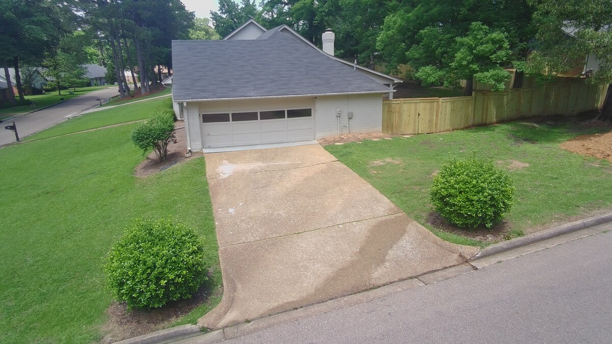 The image shows a single-story home from a vantage point, featuring a driveway leading to a two-car garage. The surrounding yard is green and well-kept, with manicured shrubs flanking the driveway. A wooden fence is visible in the background, adding privacy to the property.