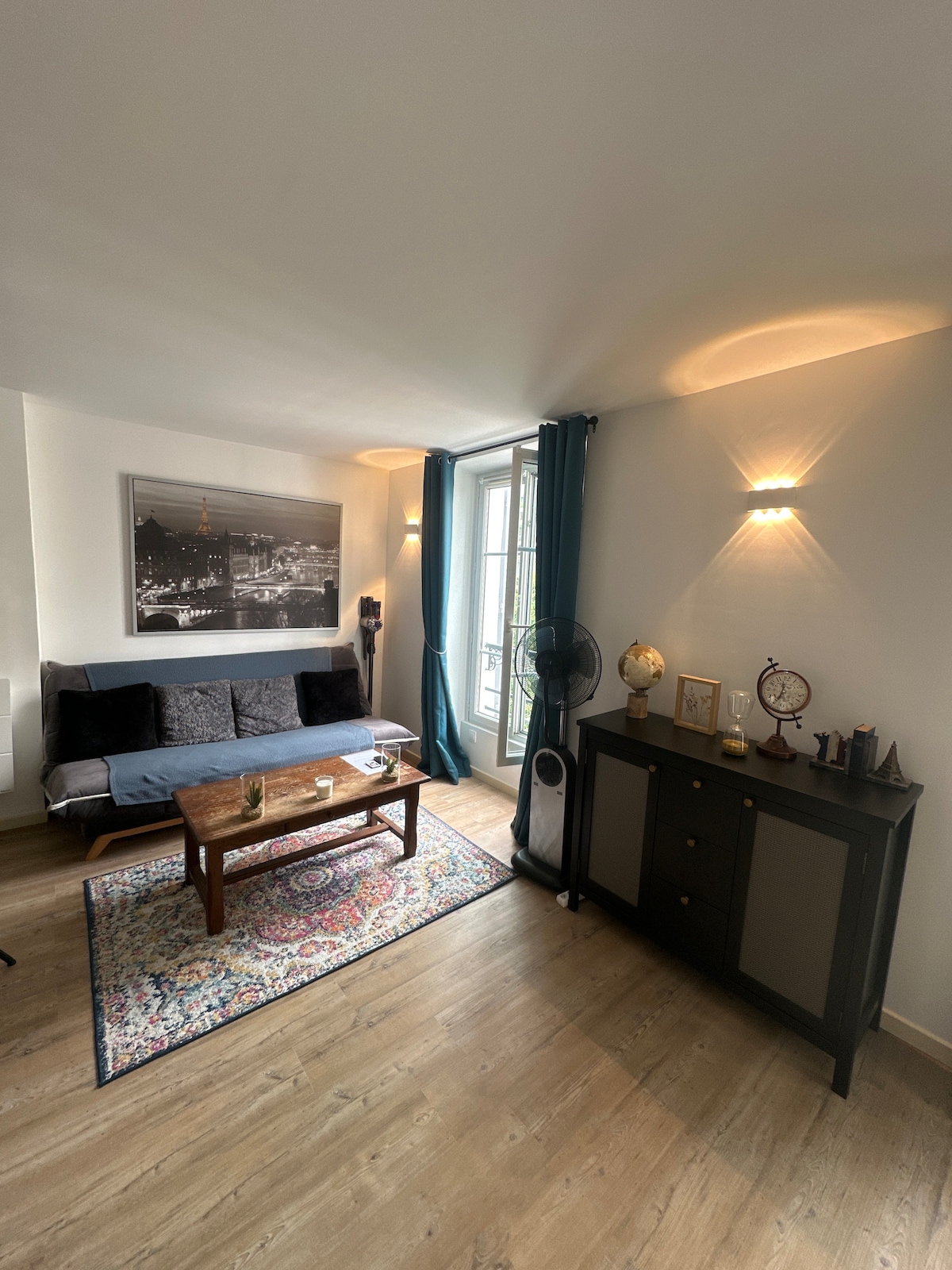 A well-lit living area features a dark sofa bed paired with a wooden coffee table, set on a patterned rug. Two curtains frame a large window, and a decorative cabinet stands nearby, complemented by wall-mounted lights that provide subtle illumination.