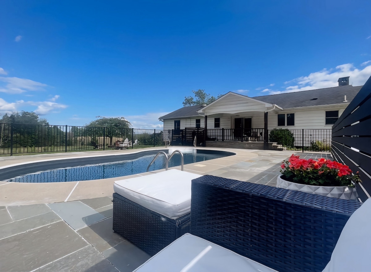 A modern home is viewed from the edge of a heated pool, surrounded by a fenced patio area. Comfortable outdoor seating is present near vibrant flower arrangements. Lush green fields and a clear blue sky provide a scenic backdrop to the tranquil setting.