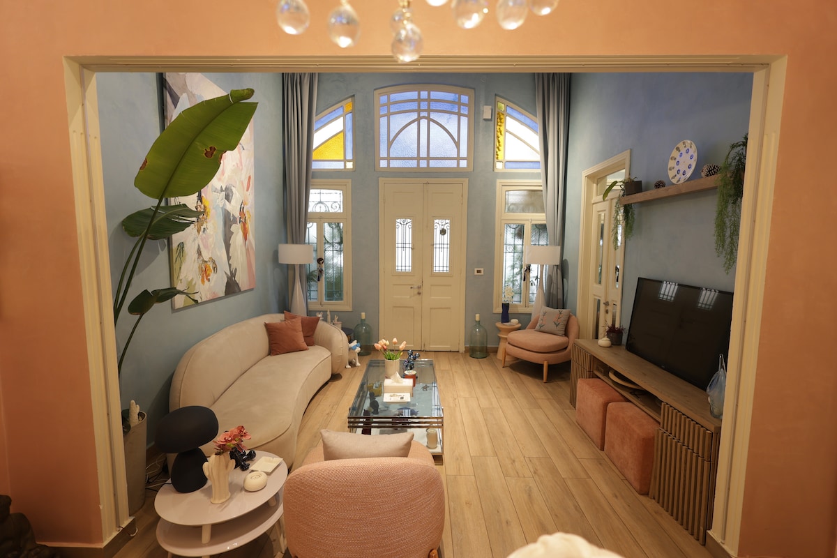 The living area features a soft beige sofa paired with light pink armchairs. Large windows allow natural light to fill the room, highlighting pastel blue walls. A wooden entertainment unit sits opposite the seating area, with decorative items arranged on top. A stylish chandelier hangs above.