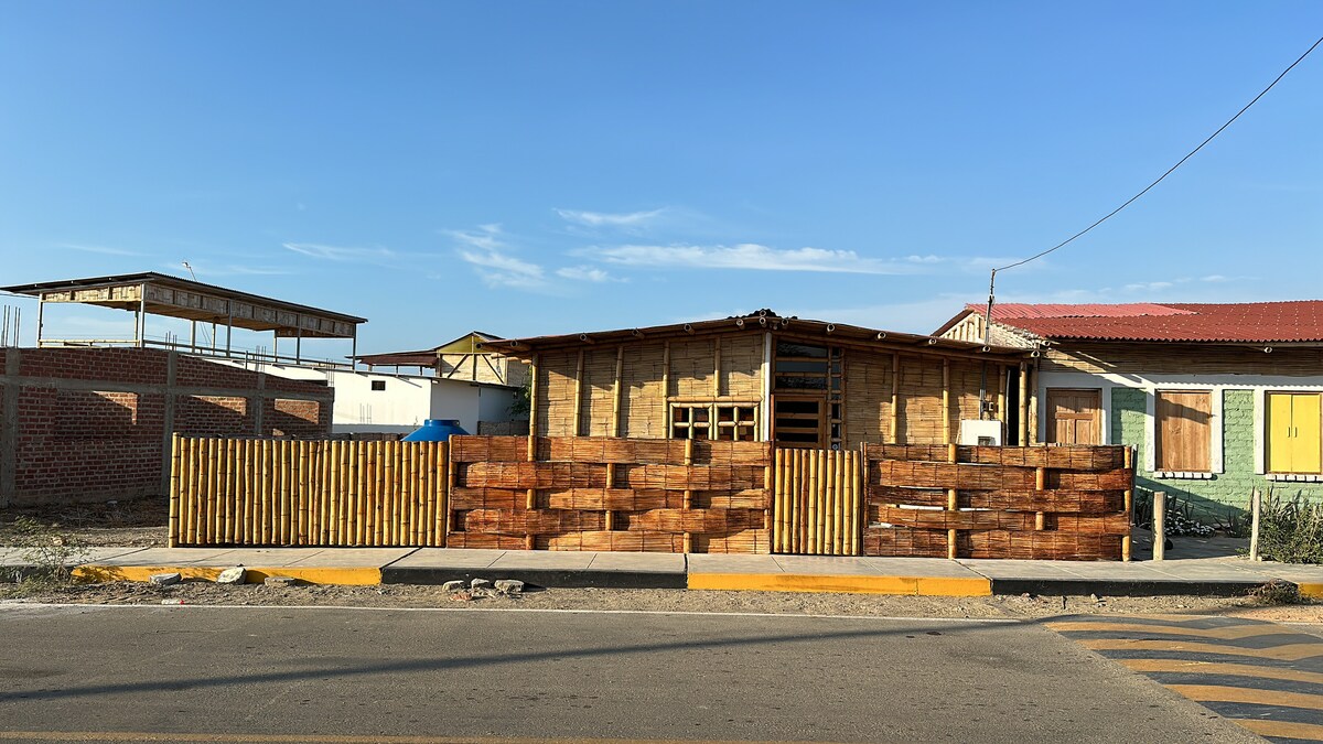 A charming bungalow is viewed from the street, featuring wooden fencing with a horizontal slat design. The main structure has a rustic feel, highlighted by large windows and a welcoming entrance. Nearby buildings and a covered area complement the space under a clear blue sky.