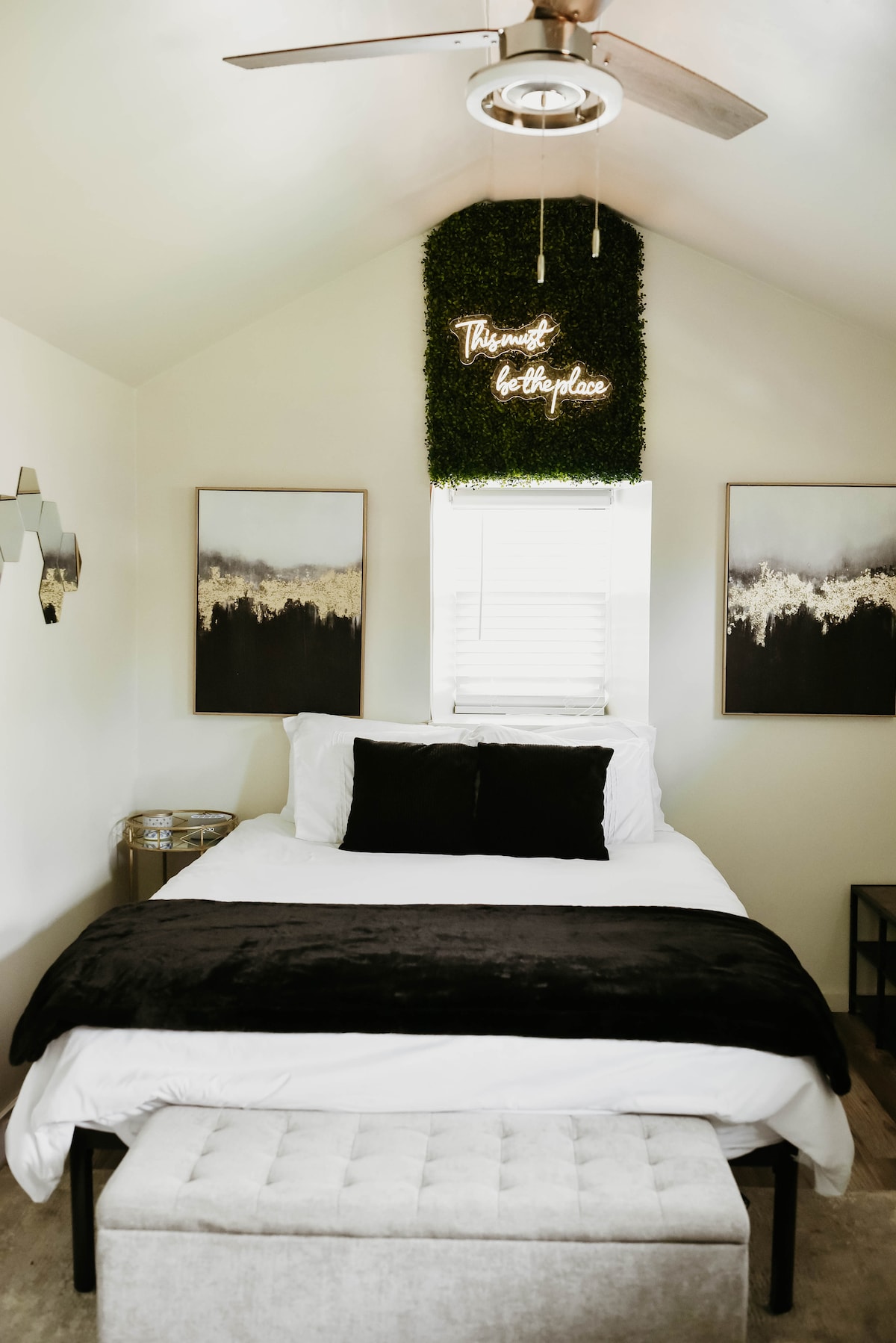 A cozy sleeping area features a stylish bed topped with a dark blanket and accent pillows. Above, a green wall decor adds a natural touch, complemented by two framed artworks. A ceiling fan is mounted above, and sunlight filters gently through the window.