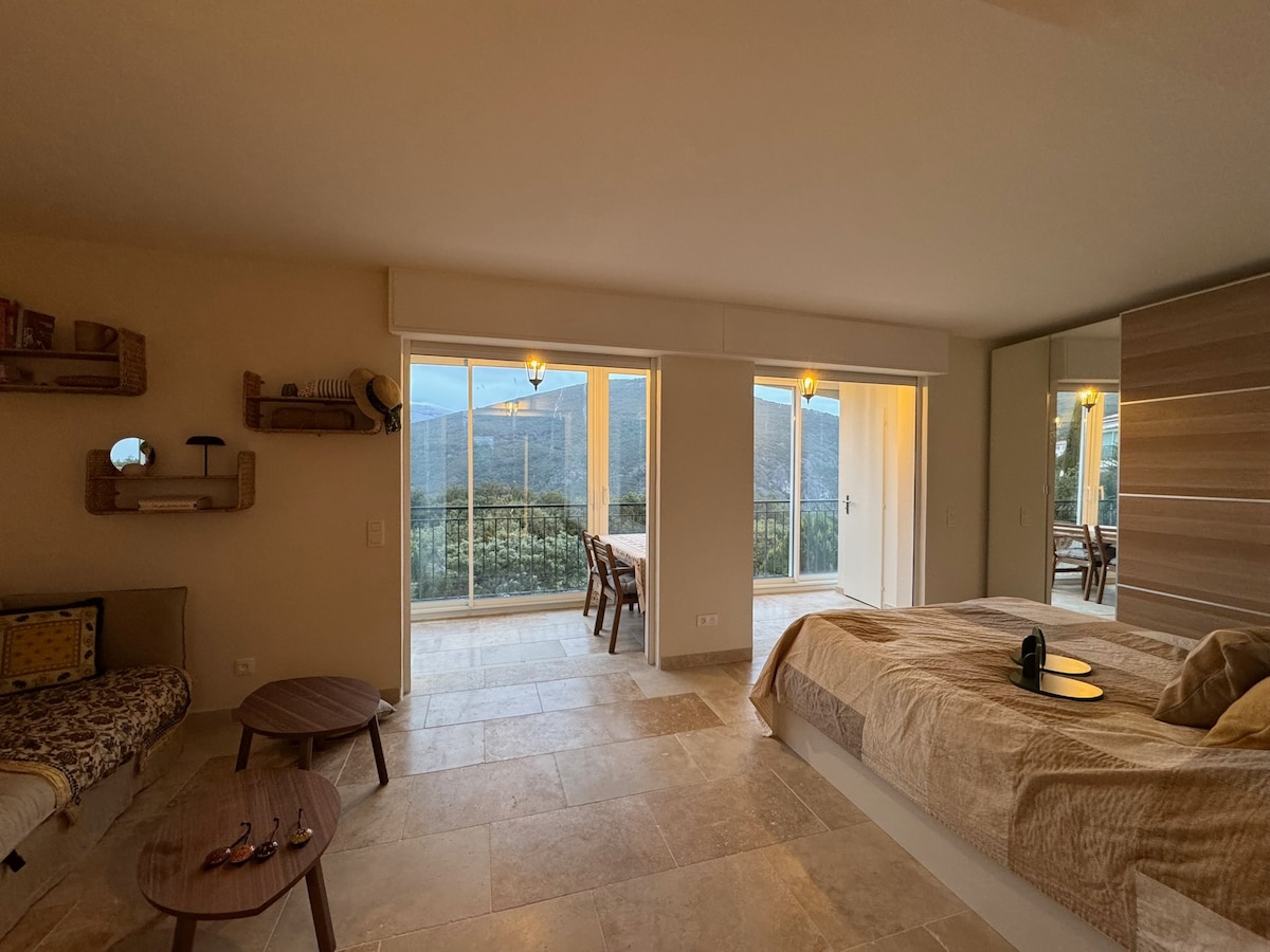 An open studio space features a spacious bed adorned with neutral linens, complemented by a wooden headboard. Natural light floods the room from large glass doors, which provide views of the mountains. A small sitting area includes a table and chairs, enhancing the inviting indoor ambiance.