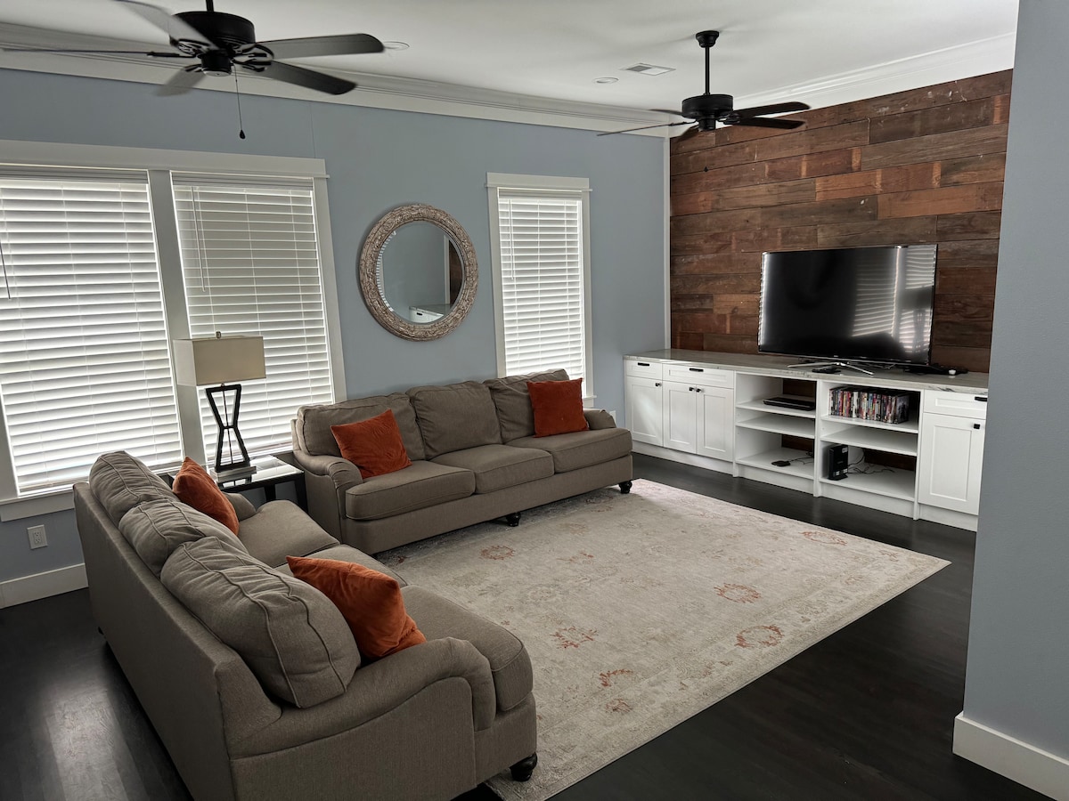 A bright living area features comfortable sofas with orange accents arranged around a central area rug. Natural light filters through multiple windows with white blinds, illuminating a wall-mounted television opposite a white media console. A decorative mirror is positioned on the wall.