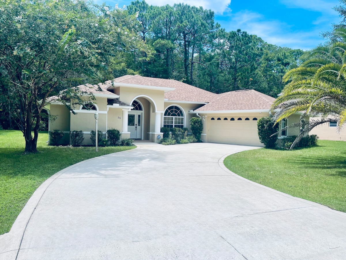 The home features a welcoming façade with a curved driveway and lush greenery surrounding it. A tiled roof and decorative arched windows enhance its charm, while a palm tree adds a tropical touch to the landscape.