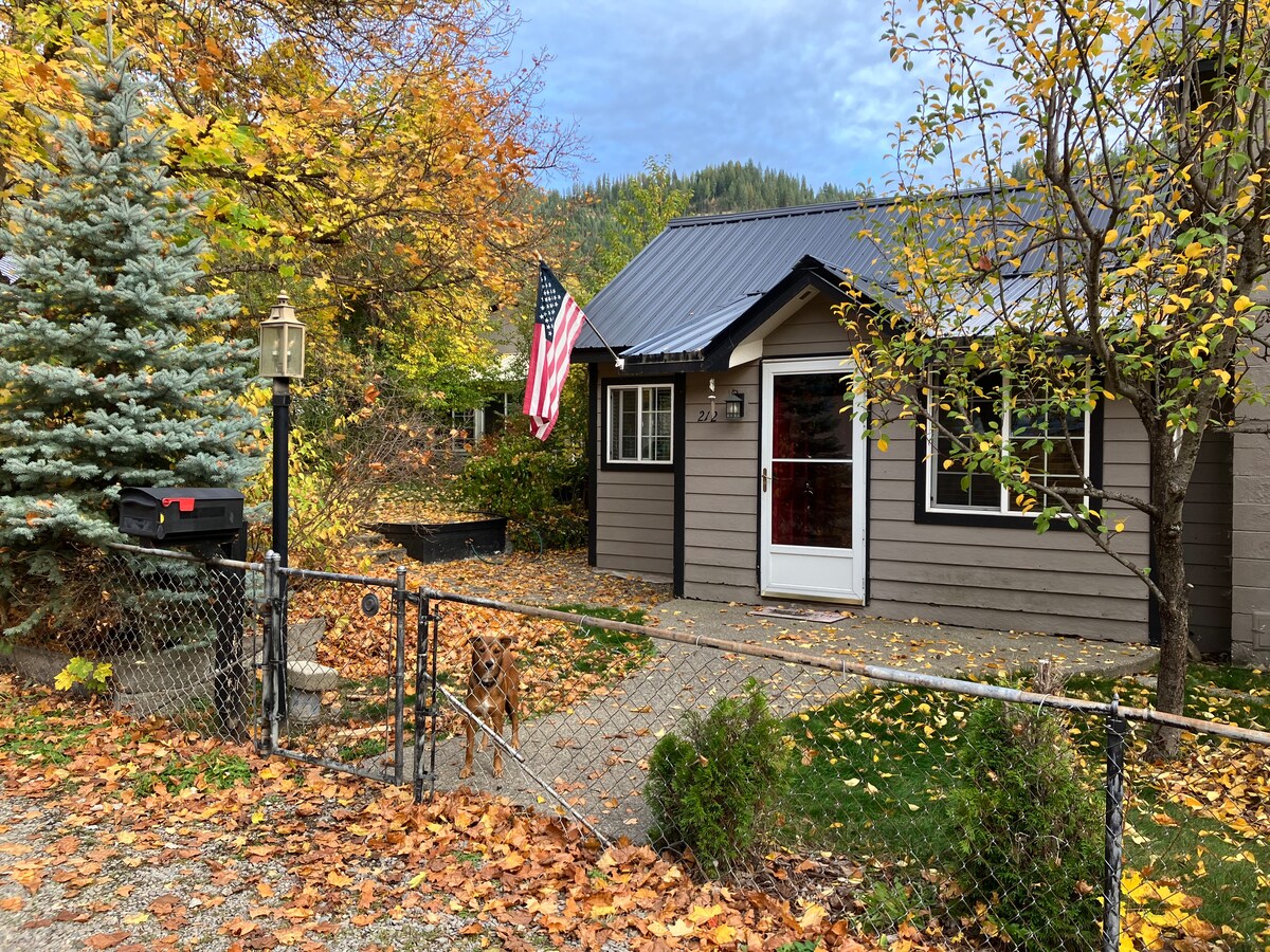 A cozy cabin is framed by vibrant autumn foliage, showcasing a well-maintained exterior with dark shingles and light gray siding. An American flag hangs from the porch, while a gravel path leads to the front entrance, flanked by decorative shrubs and trees.