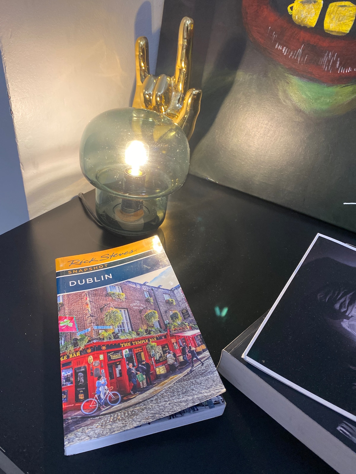 A bedside table is featured with a vibrant travel guide to Dublin, positioned alongside a stylish glass lamp with a warm light. A golden hand sculpture adds an artistic touch, while a framed photo is subtly displayed in the background.