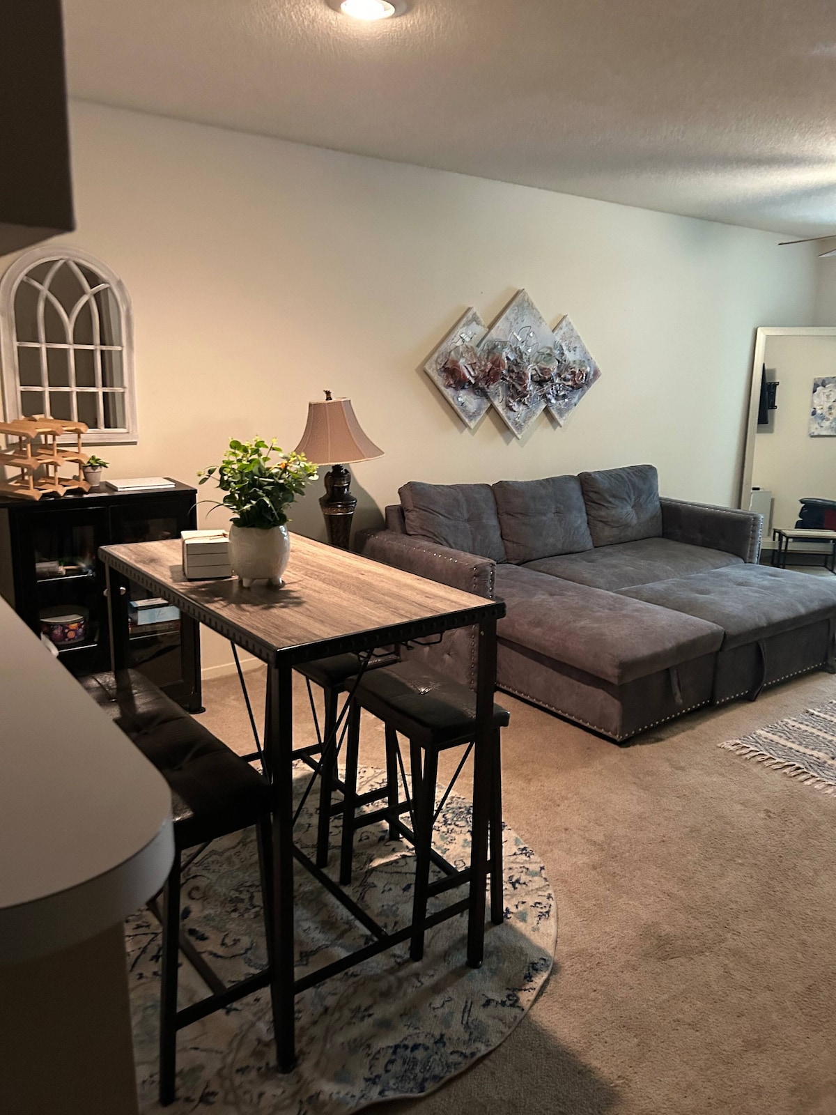 A cozy living area features a gray sectional sofa, offering casual seating. A small dining table with four stools is positioned nearby, accompanied by a decorative lamp. Subtle wall art complements the space, and a light-colored rug adds warmth to the carpeted floor.