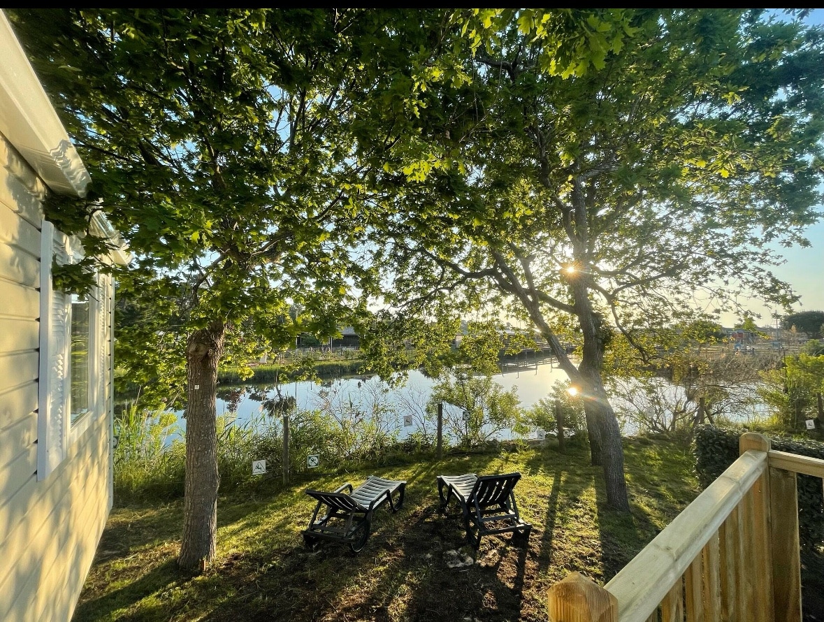 A peaceful outdoor area is shown, featuring two loungers nestled under large trees. The sun filters through the leaves, creating a dappled light effect over the lush greenery and the tranquil water of the nearby basin. A wooden railing indicates the proximity to the mobile home's terrace.