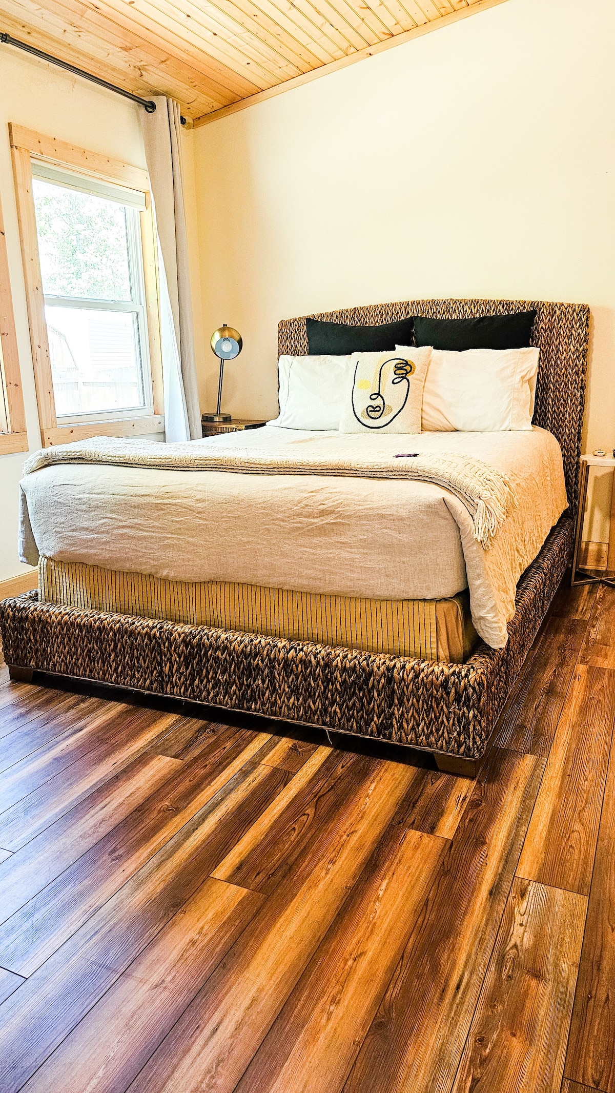 A cozy bedroom is highlighted by a spacious bed with a textured headboard and soft linens. Natural light filters in through a window adorned with light curtains. The warm wood flooring adds an inviting touch to the serene atmosphere.