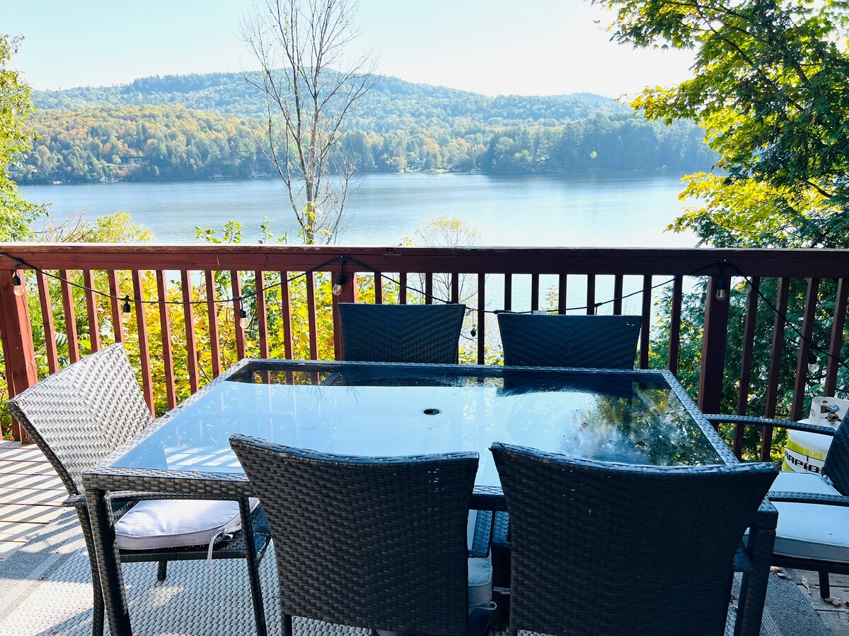 A dining table with a glass top and six wicker chairs is positioned on a deck overlooking a serene lake. Colorful autumn foliage surrounds the scene, enhancing the tranquil atmosphere with distant hills visible in the background.