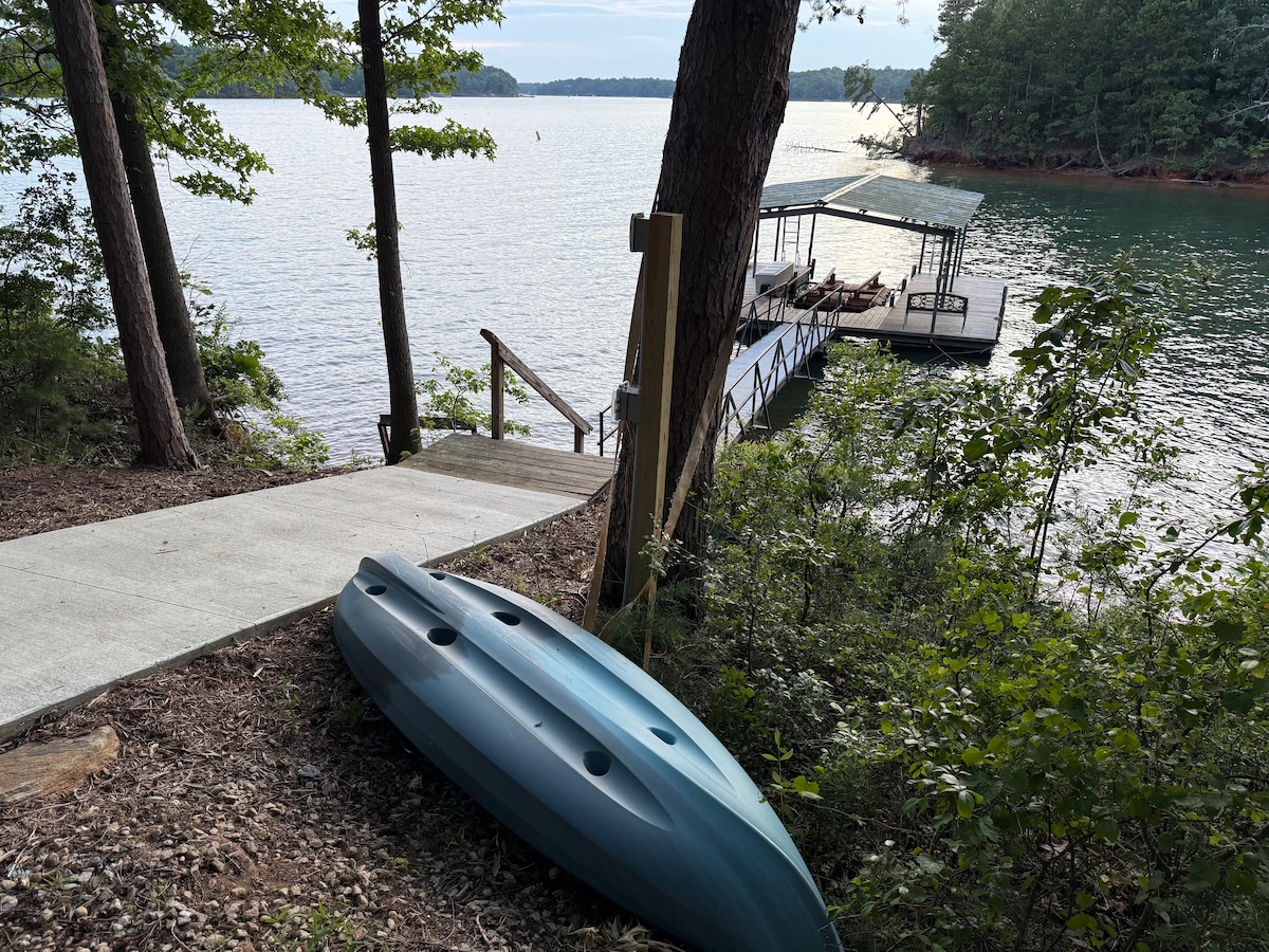 A kayak rests on the embankment beside a concrete pathway leading down to a private dock. The dock is visible over the calm waters of a lake, framed by lush trees on either side. The peaceful setting highlights a natural lakeside retreat.
