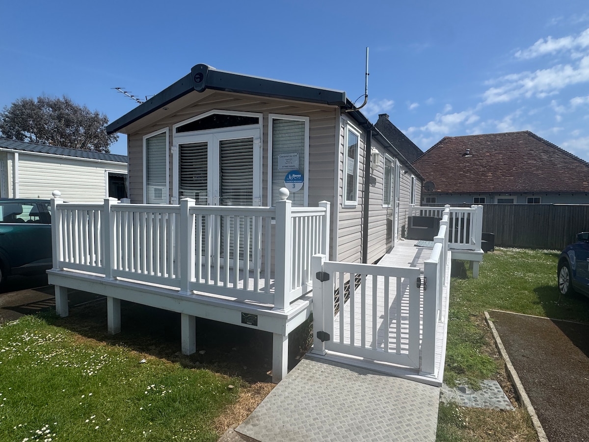 The exterior of a static caravan is featured, showcasing a spacious decking area with white railings and a ramp leading to the entrance. The bright sky and surrounding green grass create a welcoming environment, inviting guests to relax and enjoy the outdoor space.